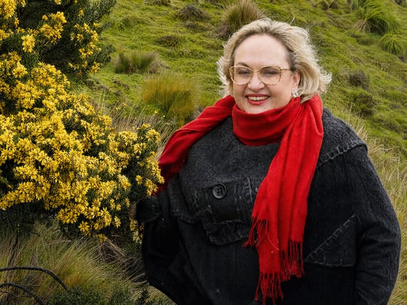 María Eugenia Puente, sonriente, con lentes, bufanda roja y chaqueta negra, al lado de una flor silvestre del páramo.