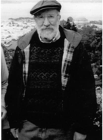 Fotografía en blanco y negro de Fernando Ortiz Crespo, con barba canosa, gafas y gorra, al aire libre con una ciudad y una bahía al fondo.