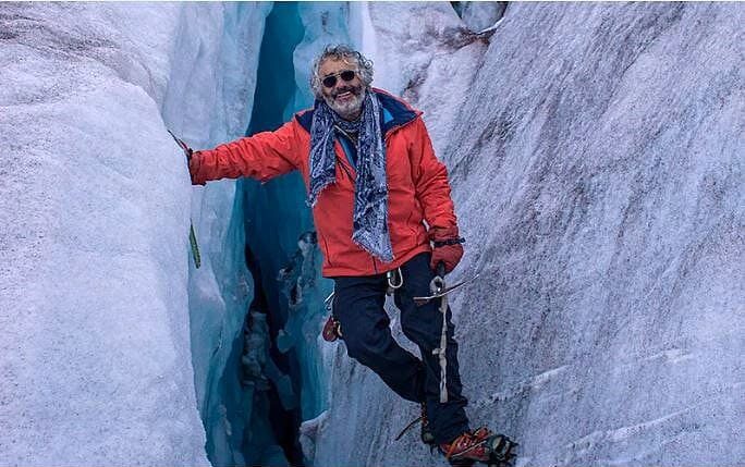 Jorge Juan Anhalzer sonríe junto a una grieta en un glaciar, con chaqueta naranja, bufanda azul, gafas de sol, crampones y piolet.