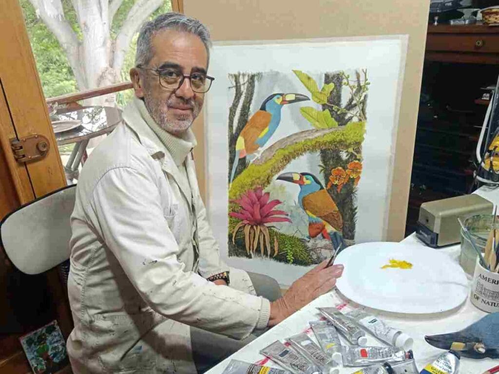 Juan Manuel Carrión sentado en su estudio, sonriente, con cabello canoso, barba corta, lentes y chaqueta beige, junto a una mesa con tubos de pintura. Detrás de él, una de sus ilustraciones de dos tucanes entre bromelias rosadas y flores anaranjadas.