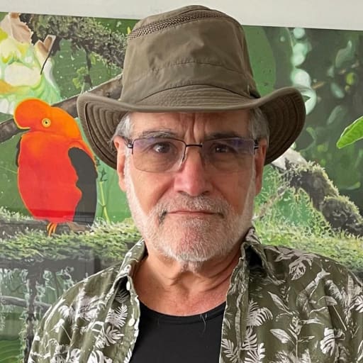 Paul J. Greenfield, con sombrero y lentes, frente a una pintura de un ave roja entre ramas cubiertas de musgo.