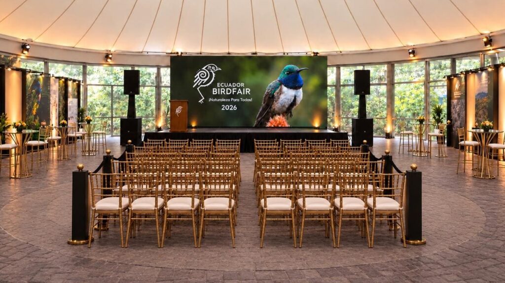 Imagen creada con IA de un auditorio con techo de carpa, sillas doradas frente a un escenario y una pantalla de Ecuador BirdFair 2026 con un colibrí.
