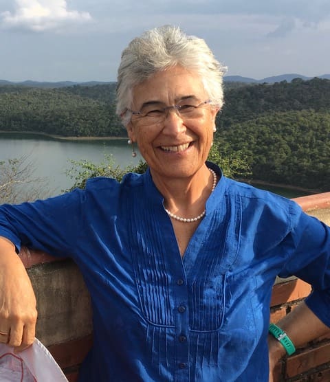 Yolanda Kakabadse, sonriente, con cabello corto y canoso, lentes y collar de perlas, blusa azul, apoyada en un muro de ladrillo frente a un lago rodeado de bosque verde.